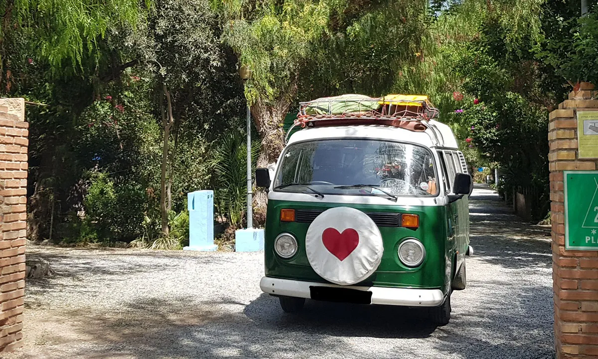 Furgoneta camper verde con un corazón pintado entrando por el camino arbolado del camping